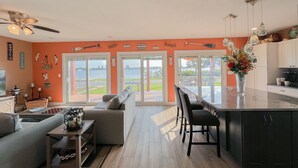 A modern living room with a sofa, a dining area with chairs, a kitchen with a bar, and large sliding glass doors leading to a view of the water.