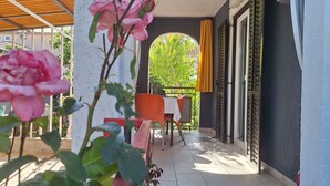 Zona relax all'aperto coperta, con tavoli e sedie, vista su un albero attraverso un arco e un edificio con persiane.