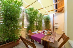 A dining area with a wooden table, chairs, and a purple tablecloth. There are potted plants and a window with blinds.