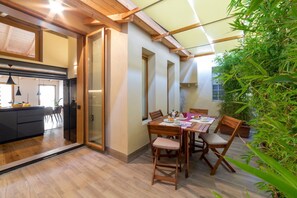 A dining area with a table and chairs, a kitchen in the background, and a large window.