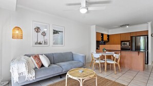 A modern living room with a grey sofa, a round wicker coffee table, and a dining area with wooden chairs. A kitchen with wooden cabinets and stainless steel appliances is visible in the background. - Seaforth Resort (Alexandra Headland)