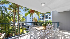 A balcony with a wooden table and chairs, a potted plant, and a view of palm trees and buildings. - Seaforth Resort (Alexandra Headland)