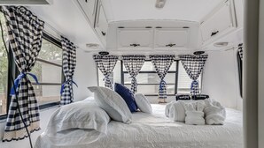 A bedroom with a bed, pillows, a small table with towels, checkered curtains, and a window with a view. - River Access, 20% Off, Glamping, Couple, Nature (New Braunfels)