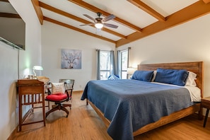 A bedroom with a large bed, wooden headboard, a desk with a chair, a red chair, a ceiling fan, and a painting on the wall.