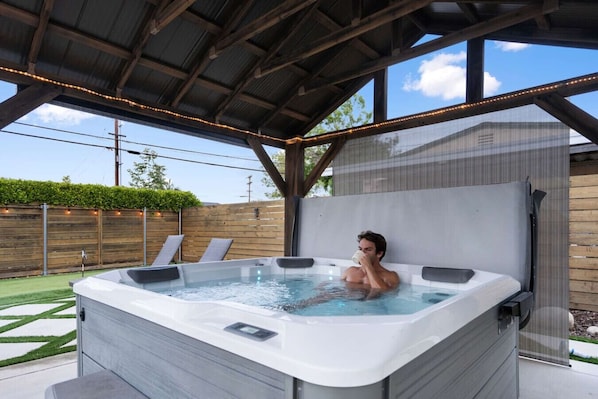 A hot tub with a person inside, surrounded by a covered patio area with a wooden fence and a grassy lawn.