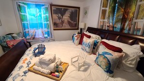 A neatly made bed with a tray of toiletries, a framed painting of a beach scene, and a window with a view of palm trees and a beach.