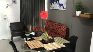 Un área de comedor con una mesa negra, cuatro sillas negras, un sofá estampado, una planta en maceta y una obra de arte colgada en la pared.