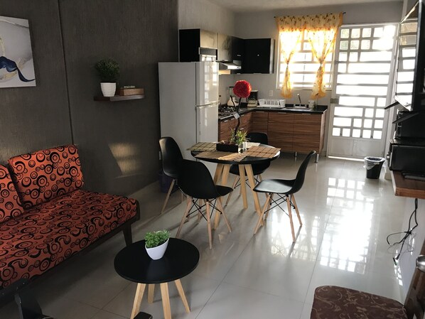 Un salon moderne comprenant un canapé, une table à manger avec des chaises, un coin cuisine avec un réfrigérateur et une fenêtre avec des rideaux.