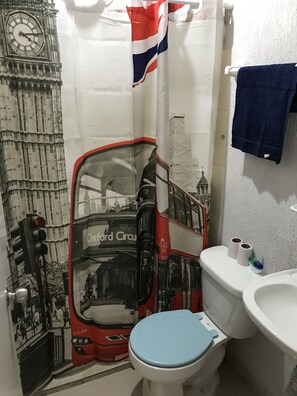 A bathroom with a toilet, sink, and a shower curtain featuring a bus and clock design. - Fully equipped apartment in the southern part of the city (San Pedro Tlaquepaque)