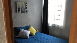 Un dormitorio con una colcha azul, una almohada blanca y una almohada amarilla. Se aprecia una ventana con cortinas, una pared con una obra de arte de motivos de hojas, y una puerta parcialmente visible.