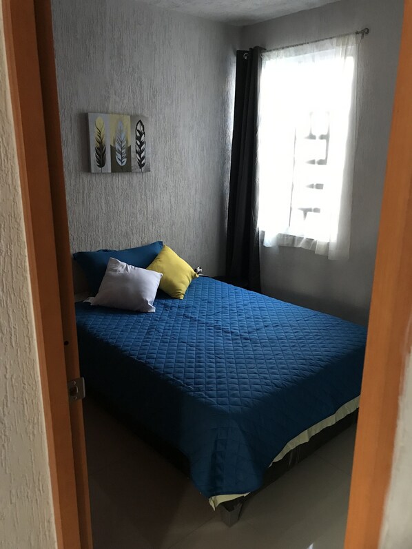 A bedroom with a blue bedspread, a white pillow, and a yellow pillow. There is a window with curtains, a wall with a leaf-patterned artwork, and a door partially visible. - Fully equipped apartment in the southern part of the city (San Pedro Tlaquepaque)