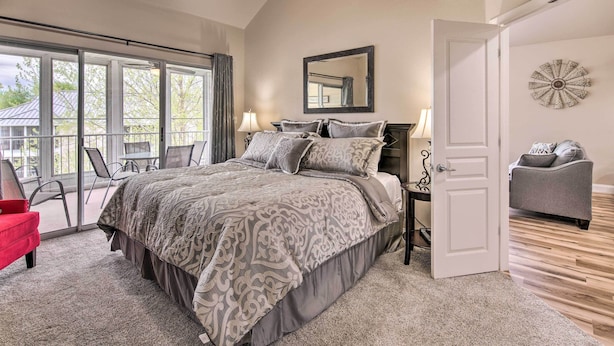 A bedroom with a large bed, a ceiling fan, a window with curtains, a bedside table with a lamp, and a door leading to another room. - Cozy + Deluxe Branson Condo w/ Covered Balcony (Branson)
