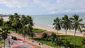 A coastal road with palm trees, a beach, and a distant city skyline. - Carpediem - Apart beira mar Cabo Branco (João Pessoa)