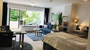 Ein modernes Hotelzimmer mit einem großen Bett, einer Sitzecke, einem Essbereich mit Tisch und Stühlen und Blick ins Freie durch große Fenster.
