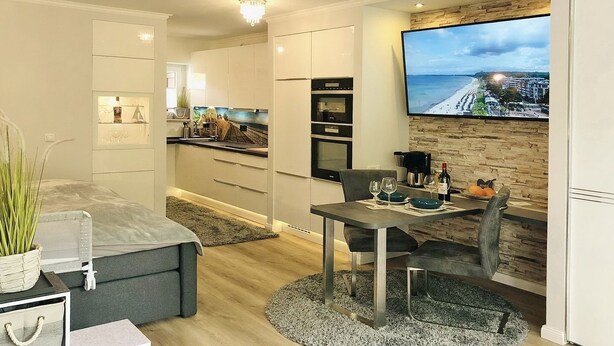 Ein modernes, kompaktes Wohnzimmer mit Küchenzeile, einem Essbereich mit Tisch für zwei Personen und einem großen Fernseher an der Wand, der eine Strandansicht zeigt.
