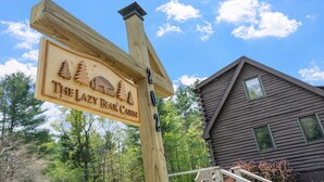 Un poste de madera con la inscripción 'The Lazy Bear Cabin', una cabaña de madera y un cielo azul despejado con nubes.