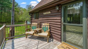 Una cabaña de troncos con una terraza de madera, dos sillas Adirondack, un cartel de bienvenida y un ventanal de vidrio.
