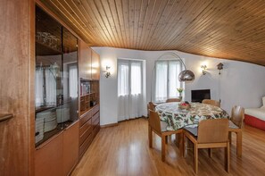 Un coin repas doté d’un plafond en bois, d’une table ronde entourée de chaises, d’un téléviseur et d’une bibliothèque où sont exposés des objets en verre.