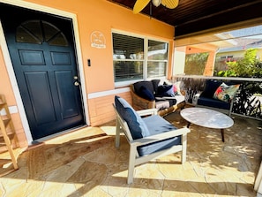 A covered patio area with a black door, a wicker sofa, a white armchair, and a round table.