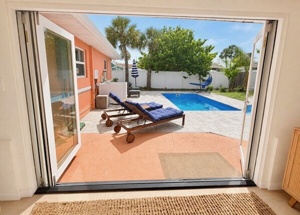 Vista a través de una puerta de cristal que da a un patio trasero con piscina, dos sillones de descanso y una pequeña zona de patio.