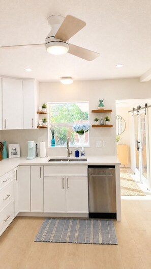 A modern kitchen with white cabinets, a stainless steel dishwasher, a ceiling fan, and a window with a view of greenery. -  Tropical Beach Bungalow w/Private Saltwater Pool and Additional Casita (St. Pete Beach)