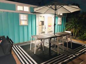 Un espace extérieur aménagé pour les repas, avec une table et des chaises, un tapis à rayures et un grand parasol.