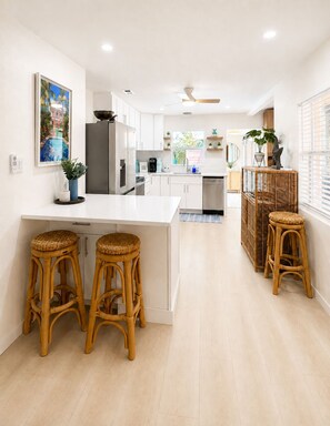 Una cocina moderna con muebles blancos, una isla central con taburetes, un refrigerador y un comedor con una silla de mimbre.