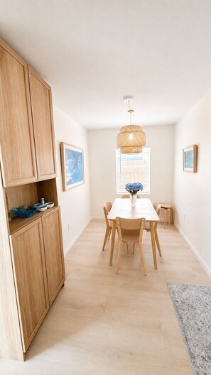 A dining area with a wooden table, chairs, a hanging light fixture, and a window with blinds.
