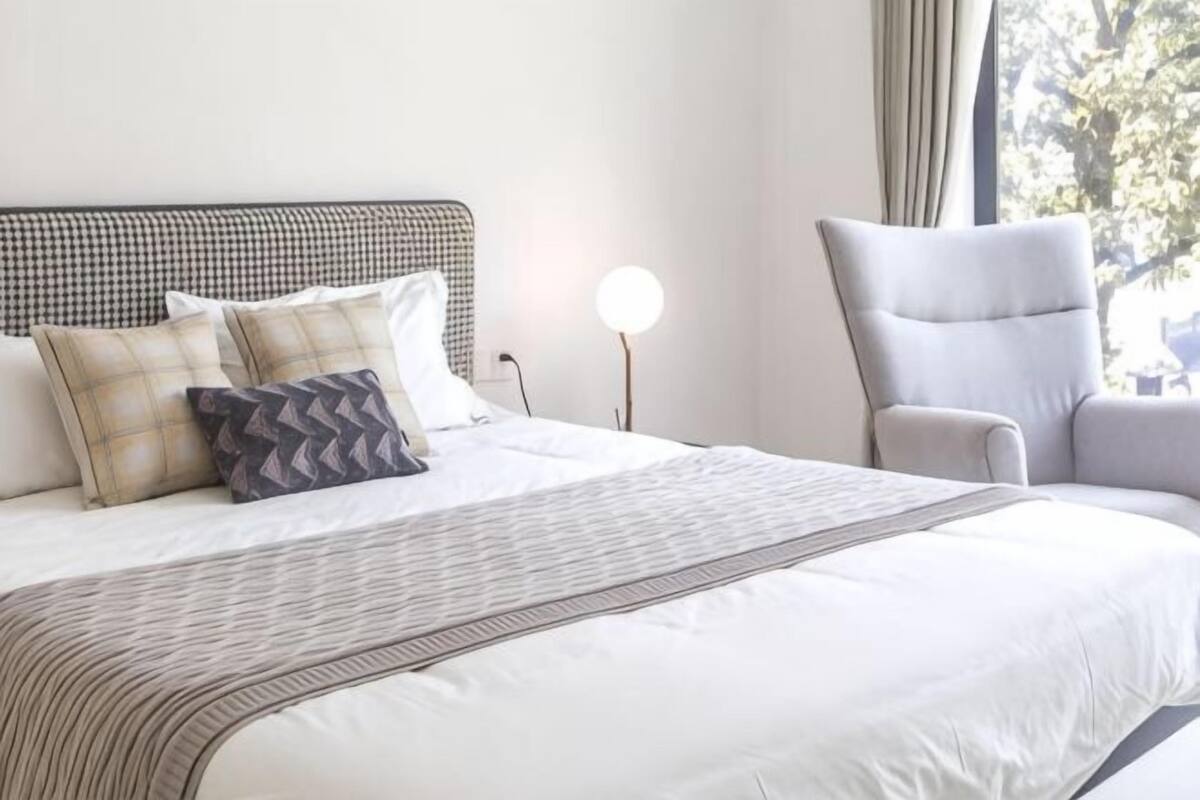 A neatly made bed with a striped bedspread, a headboard with a checkered pattern, and a chair with a light-colored cushion.