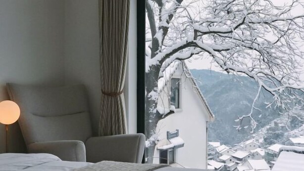 Ein Schlafzimmer mit Bett, Sessel, Lampe und Fenster mit Blick auf einen verschneiten Berg.