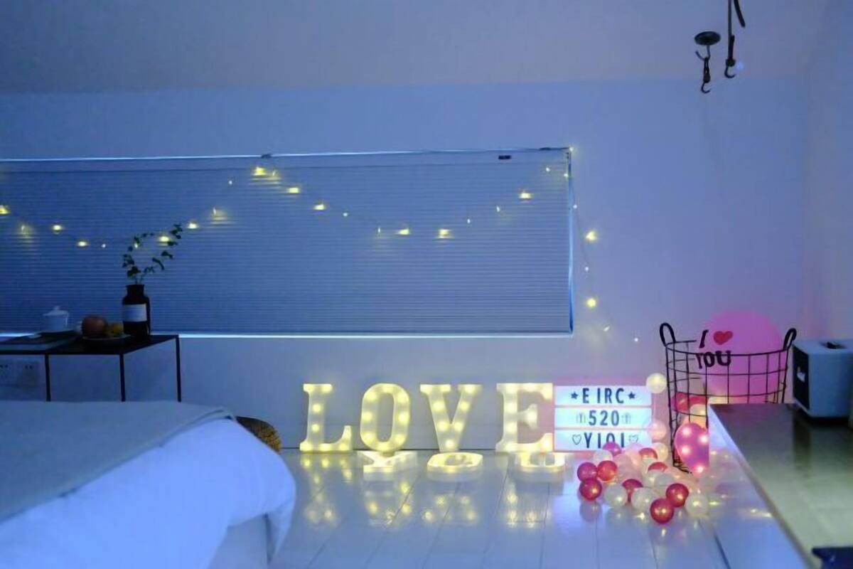 A room with a bed, a desk with a lamp, a wall with illuminated letters spelling 'LOVE', and a window with blinds.