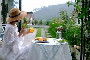 Una persona sentada a una mesa con comida y bebida, al aire libre, con un jardín y un edificio de fondo.