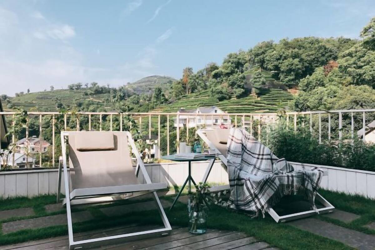 A deck with two chairs and a table, overlooking a lush green landscape with terraced fields and a house in the background.