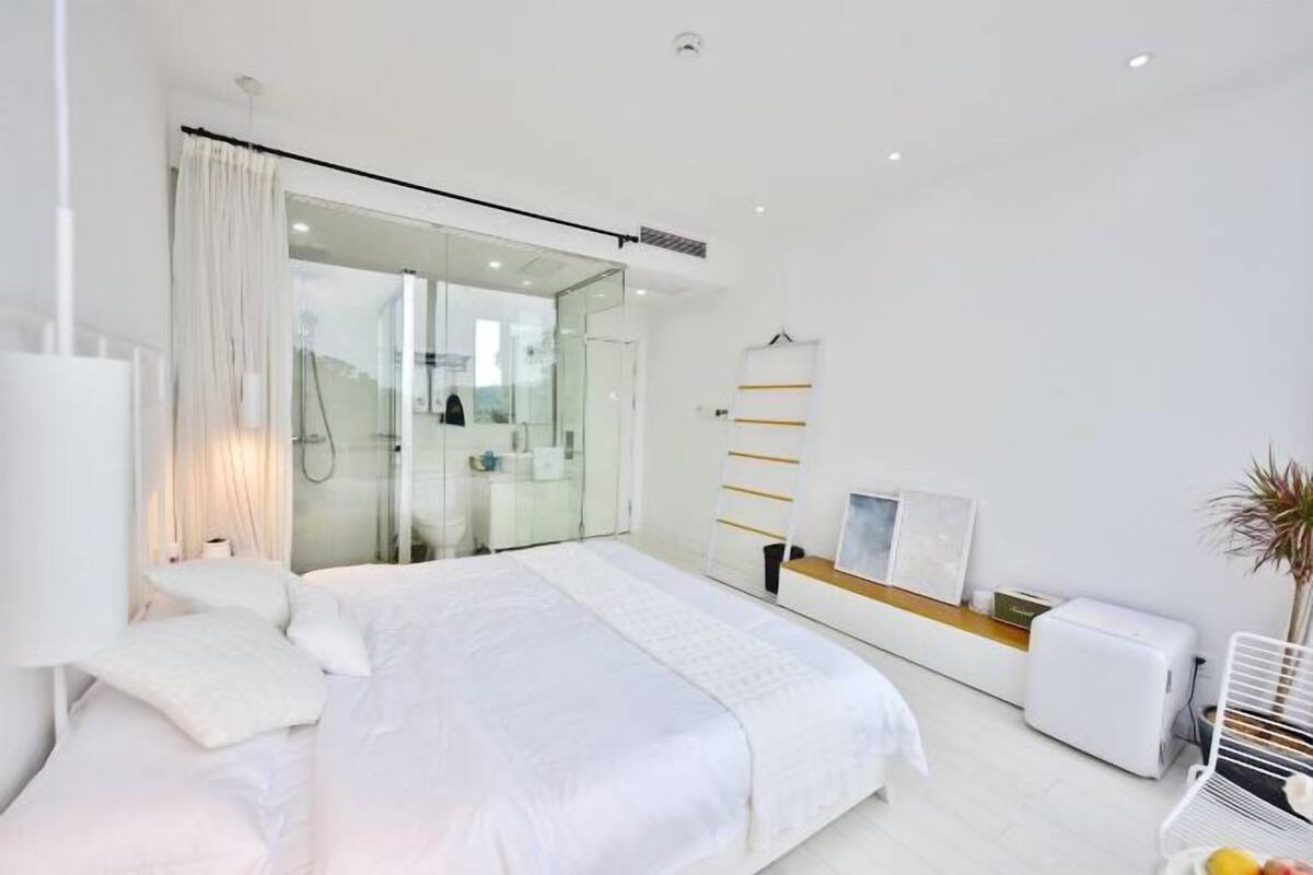 A modern bedroom with a large bed, a glass shower enclosure, a bedside table, and a small seating area.