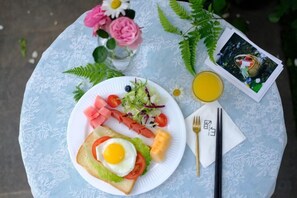 Une table ronde recouverte d’une nappe en dentelle blanche, une assiette de nourriture, un verre de jus de fruits, un cadre photo et une paire de baguettes.