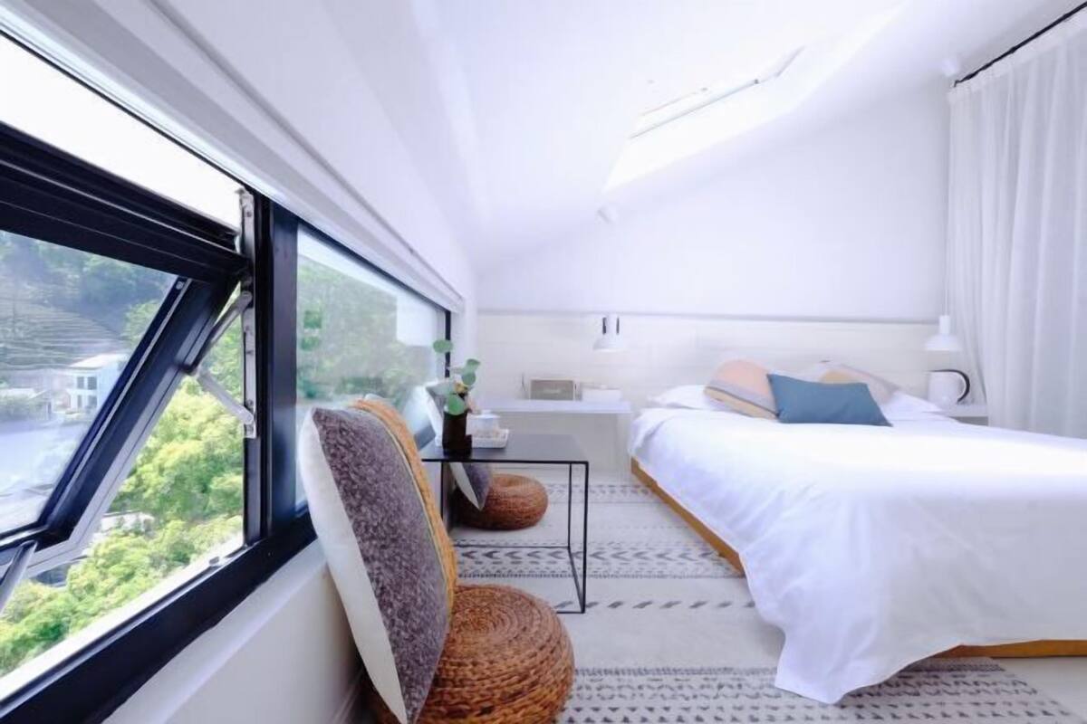 A modern hotel room with a large window, a bed, a bedside table, and a chair.