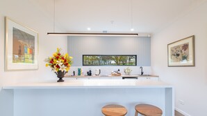 Una cocina moderna con encimera blanca, dos taburetes de madera, un jarrón de flores y una ventana con vistas a árboles.