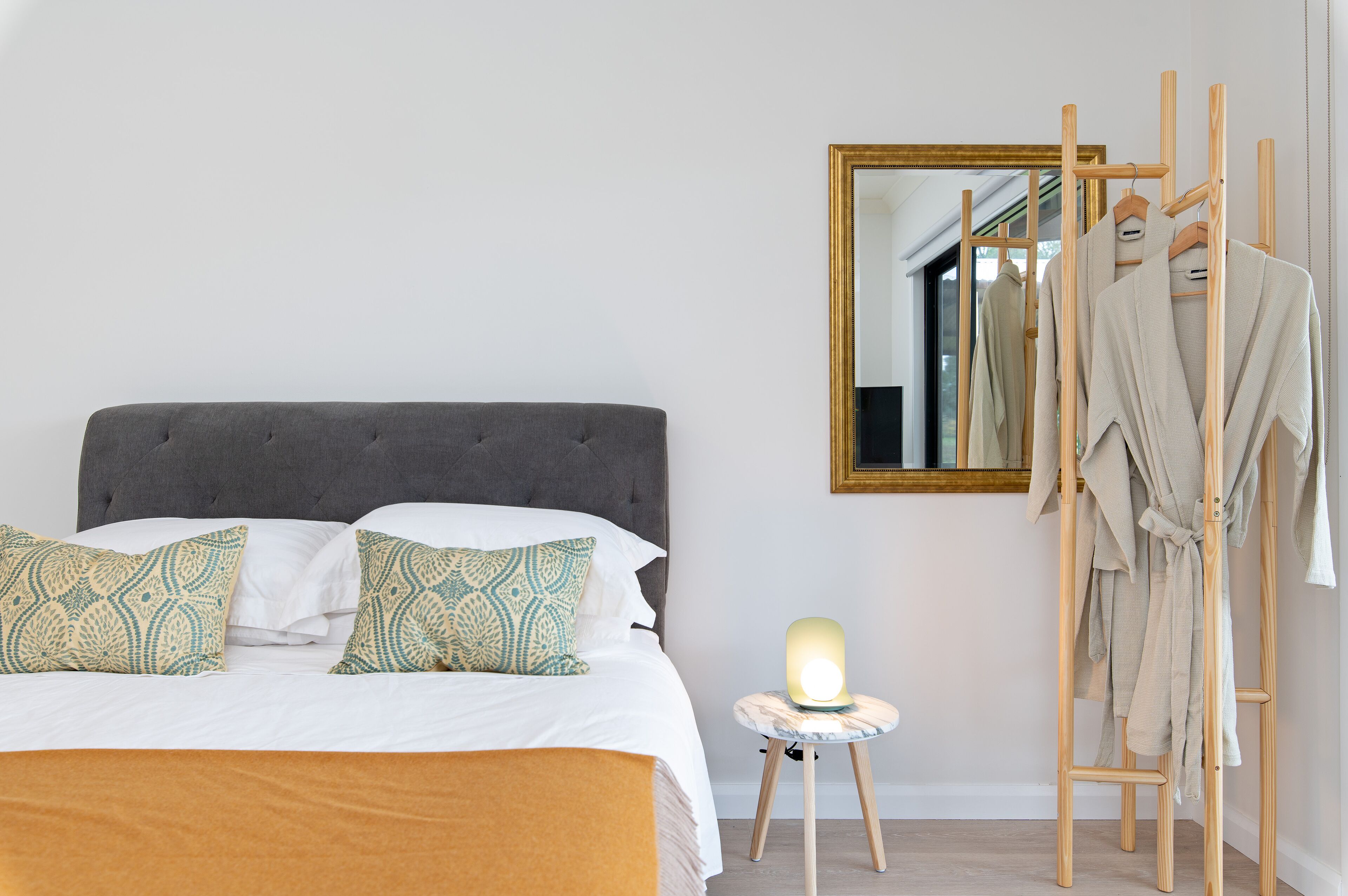 A neatly made bed with patterned pillows, a bedside table with a lamp, and a wooden robe rack against a wall with a mirror.