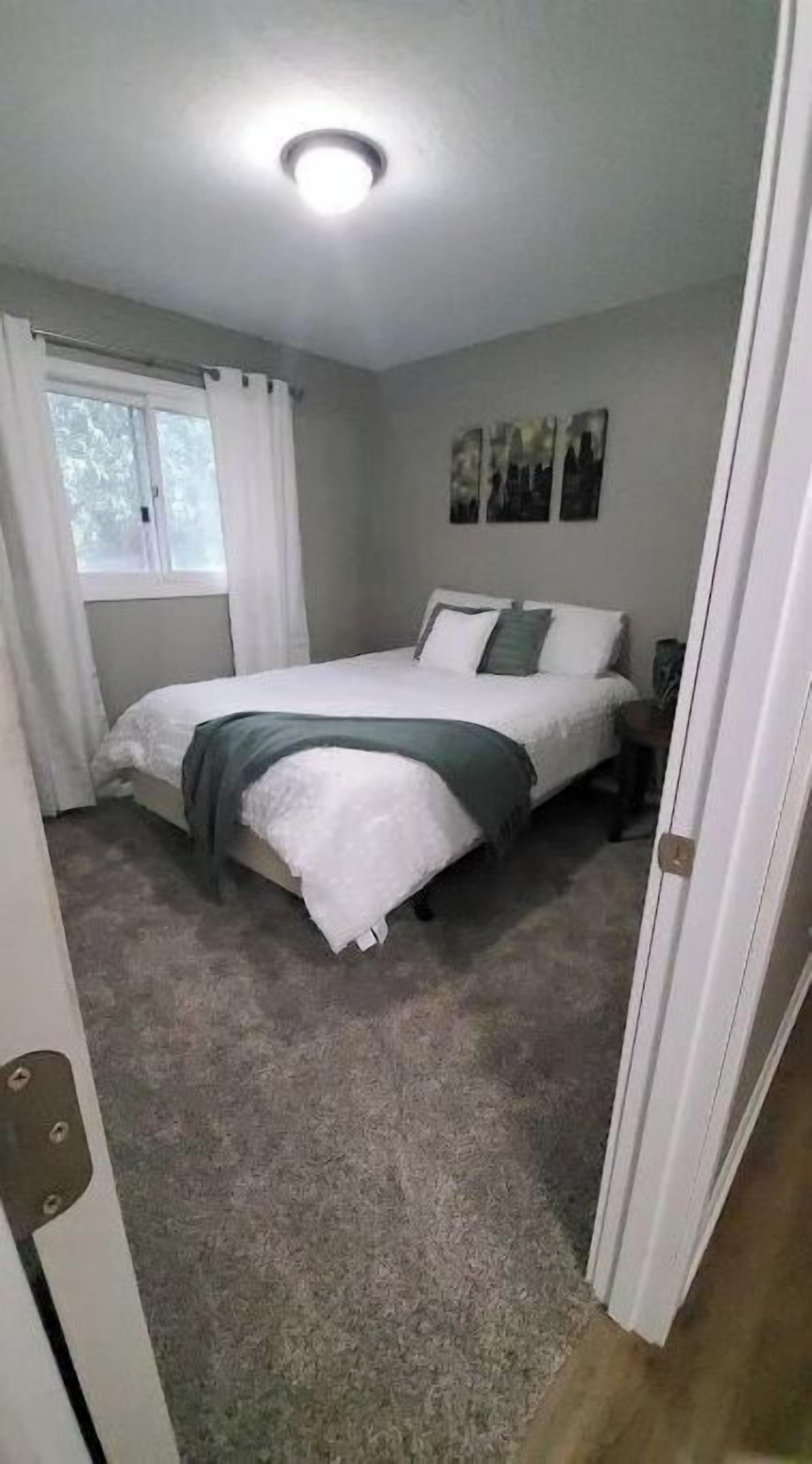 Um quarto com cama, janela com cortinas, mesa de cabeceira e uma obra de arte na parede.