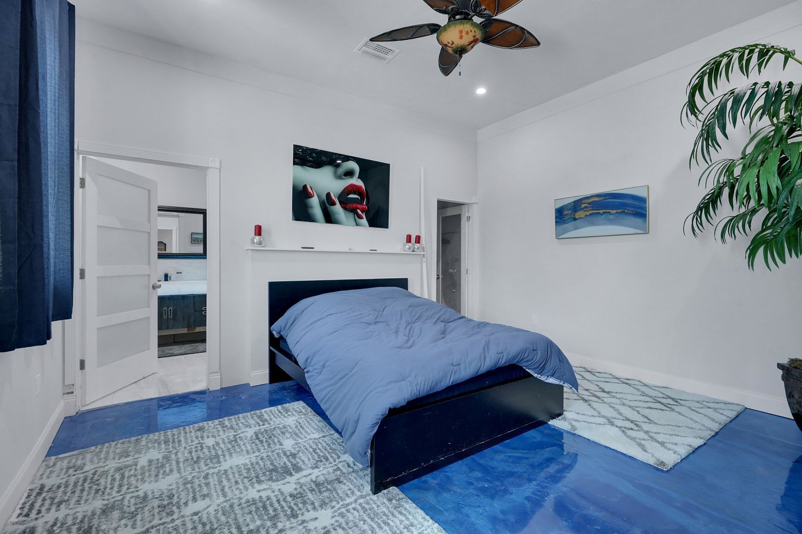 A bedroom with a bed, a ceiling fan, a TV, a plant, and a painting.