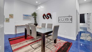 A modern dining room with a glass table, six chairs, a red and black striped rug, and wall art including a plant and abstract designs.