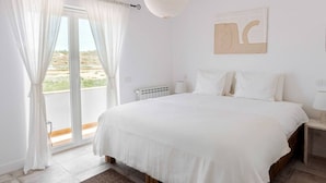 Un dormitori amb un llit gran, cortines blanques, vista a l'exterior, un radiador i un llum penjant.