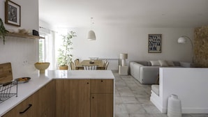 Una cuina moderna amb armaris de fusta, un taulell blanc i una zona de menjador amb taula i cadires. Hi ha un sofà i una làmpada a la zona de sala d'estar.