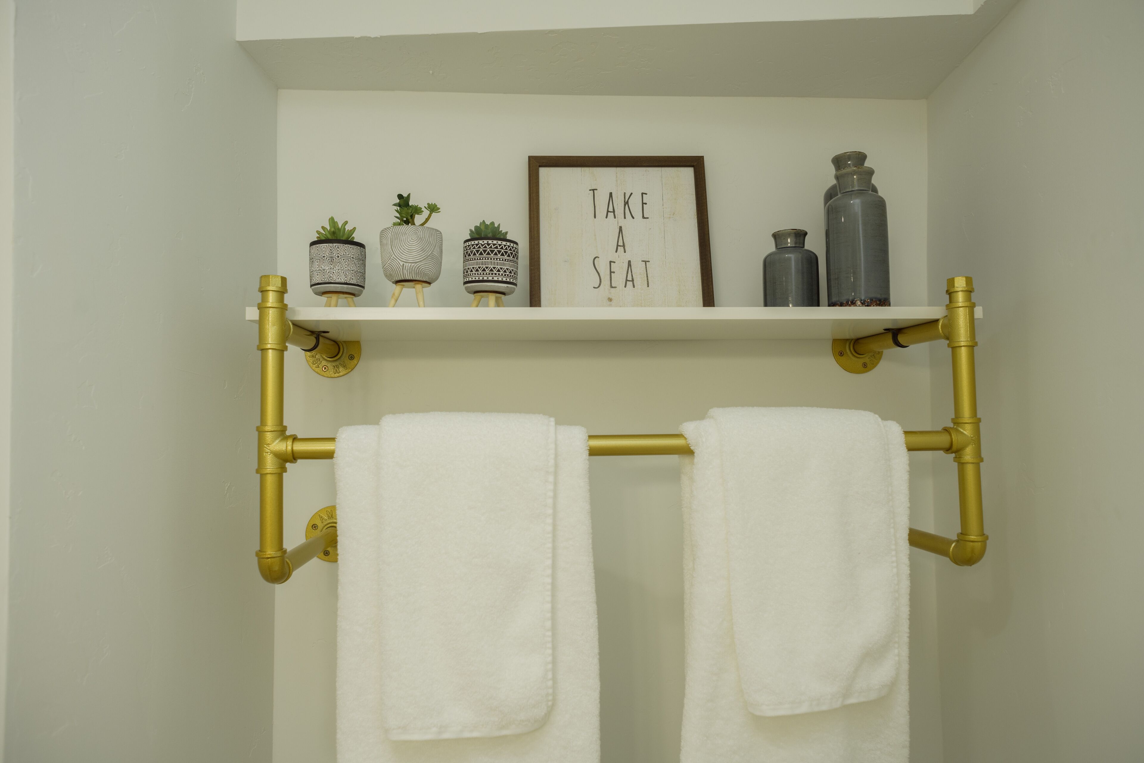 Un baño con un toallero hecho de cañerías doradas, un estante con adornos y un cartel que dice 'TAKE A SEAT'.