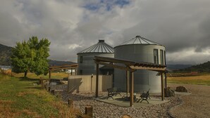 Um edifício rústico com dois silos cilíndricos, uma pérgula de madeira e um caminho de cascalho com bancos.