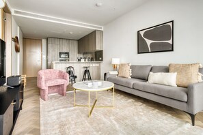 Uma sala de estar moderna com um sofá cinza, uma poltrona rosa e uma mesa de centro redonda. Uma cozinha é visível ao fundo.
