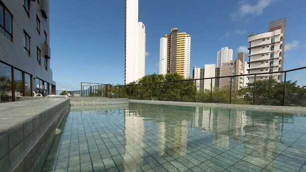 Una piscina al aire libre con suelo de baldosas, rodeada por una valla baja y árboles.