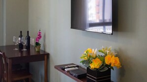Una habitación con una televisión de pantalla plana en la pared, una mesa de madera con botellas de vino y copas, y una mesita con un control remoto y un jarrón decorativo con flores.