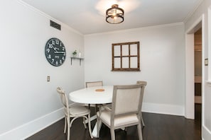 Un coin repas comprenant une table ronde et quatre chaises, une horloge murale, une fenêtre avec un cadre en bois et un luminaire au plafond.