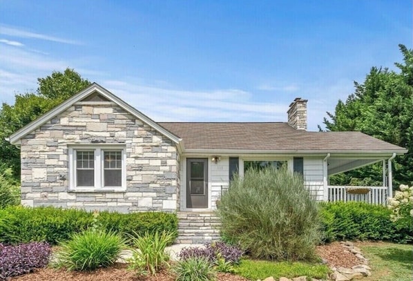 Une maison en pierre avec un porche à l’avant, une cheminée et un jardin bien entretenu.